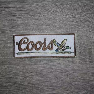 Pode incluir: Patch bordado com a palavra "Coors" em escrita castanha e uma ilustração de um pato a voar. O patch tem um fundo branco com uma borda castanha e uma forma retangular.