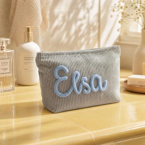 May include: A gray corduroy cosmetic bag with the name "Elsa" in light blue script. The bag is rectangular and sits on a yellow surface. Other toiletries are in the background.