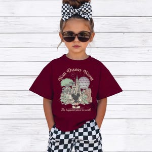 May include: A maroon t-shirt with a graphic of the Walt Disney World castle, Mickey Mouse, and the text "Walt Disney World" and "The happiest place on earth." The shirt is worn with black and white checkered shorts.