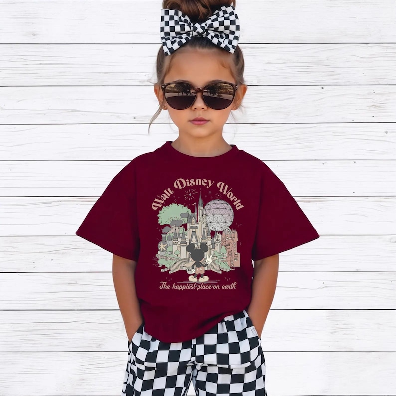 May include: A maroon t-shirt with a graphic of the Walt Disney World castle, Mickey Mouse, and the text "Walt Disney World" and "The happiest place on earth." The shirt is worn with black and white checkered shorts.