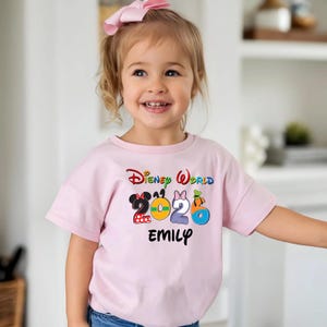 May include: A light pink t-shirt with the words "Disney World 2026" in colorful lettering. The number 2 is designed with a Minnie Mouse theme, 0 with a yellow circle, 2 with a Minnie Mouse theme, and 6 with a Donald Duck theme. The name "EMILY" is printed below.