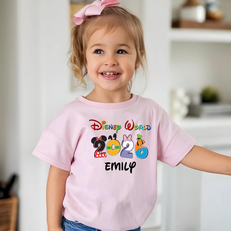 May include: A light pink t-shirt with the words "Disney World 2026" in colorful lettering. The number 2 is designed with a Minnie Mouse theme, 0 with a yellow circle, 2 with a Minnie Mouse theme, and 6 with a Donald Duck theme. The name "EMILY" is printed below.