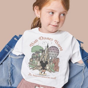 May include: White t-shirt featuring a vintage-style illustration of Walt Disney World with Mickey Mouse. The shirt has the text "Walt Disney World" and "The happiest place on earth". The shirt is worn by a young girl.