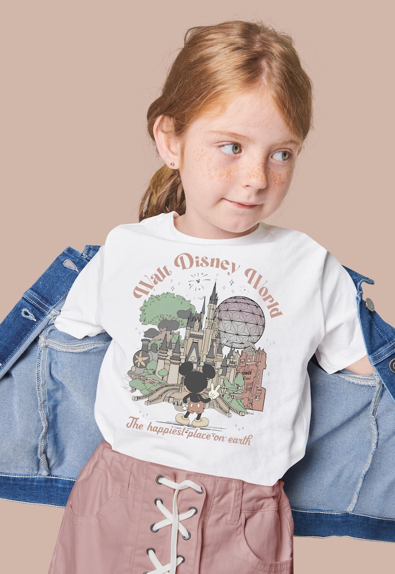 May include: White t-shirt featuring a vintage-style illustration of Walt Disney World with Mickey Mouse. The shirt has the text "Walt Disney World" and "The happiest place on earth". The shirt is worn by a young girl.