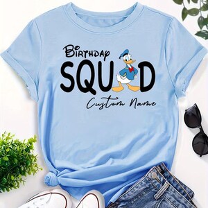 Pode incluir: Uma t-shirt de manga curta azul-clara com o texto preto "BIRTHDAY SQUAD", um gr&aacute;fico do Pato Donald a substituir a letra "A" e as palavras "Custom Name" impressas em letra cursiva preta por baixo do texto principal.