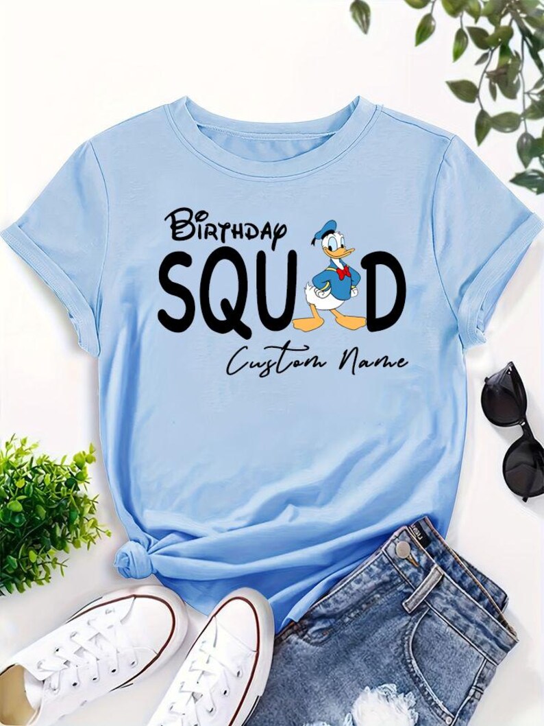 Pode incluir: Uma t-shirt de manga curta azul-clara com o texto preto "BIRTHDAY SQUAD", um gr&aacute;fico do Pato Donald a substituir a letra "A" e as palavras "Custom Name" impressas em letra cursiva preta por baixo do texto principal.