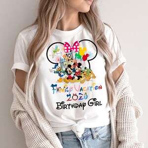 May include: A white short-sleeve t-shirt featuring a graphic of Disney characters including Mickey Mouse, Minnie Mouse, Donald Duck, Goofy, Pluto, and Tinker Bell. The text reads Family Vacation 2020 Birthday Girl with a castle and balloons.