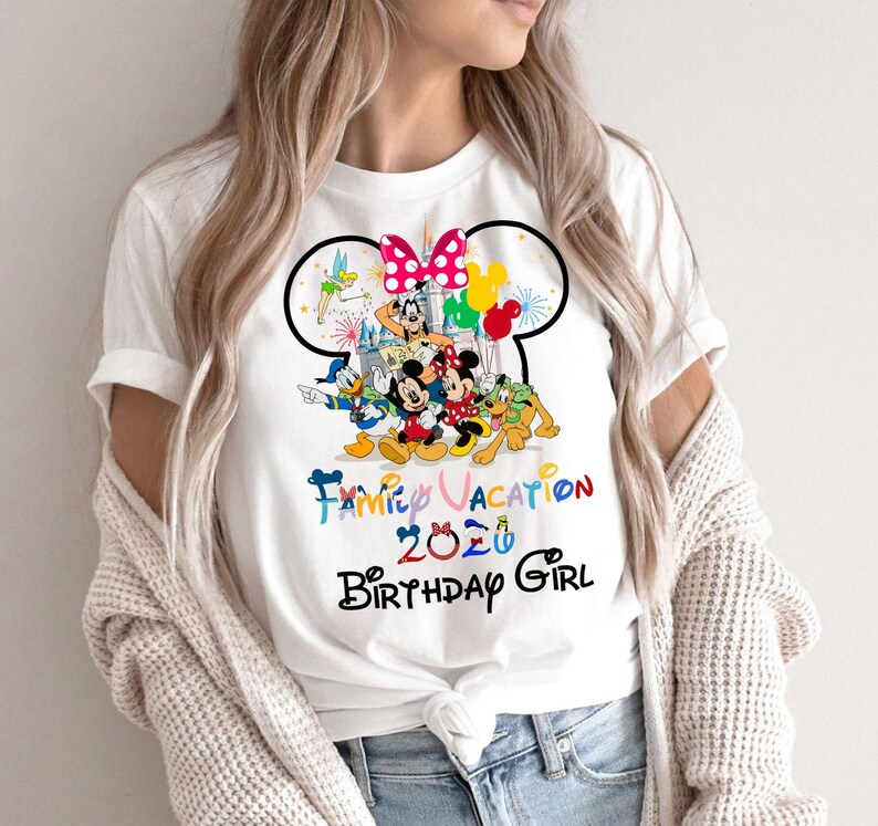 May include: A white short-sleeve t-shirt featuring a graphic of Disney characters including Mickey Mouse, Minnie Mouse, Donald Duck, Goofy, Pluto, and Tinker Bell. The text reads Family Vacation 2020 Birthday Girl with a castle and balloons.