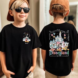 May include: A black t-shirt featuring a small graphic of Mickey Mouse and a castle on the front chest with the name Aaron, and a large graphic on the back showing Mickey, Minnie, Donald, Daisy, Goofy, and Pluto in front of a castle with Disneyland Est. 1955 text.