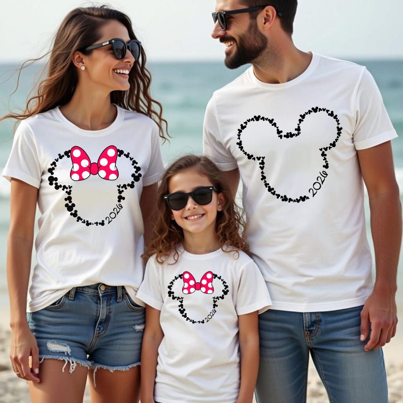 May include: Three white short-sleeved t-shirts featuring black outlines of Mickey Mouse heads composed of smaller icons. Two shirts display a pink and white polka-dot bow on the head, and all three shirts include the year 2026 printed in black.