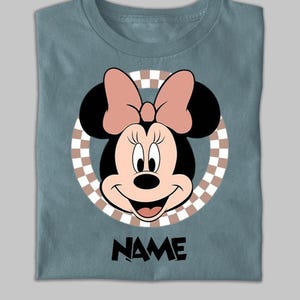 Puede incluir: Una camiseta azul pizarra con un gr&aacute;fico de Minnie Mouse centrado dentro de un c&iacute;rculo a cuadros beige y blanco. Minnie Mouse aparece con un lazo rosa, orejas negras y una sonrisa. La palabra NAME est&aacute; impresa en negro debajo.