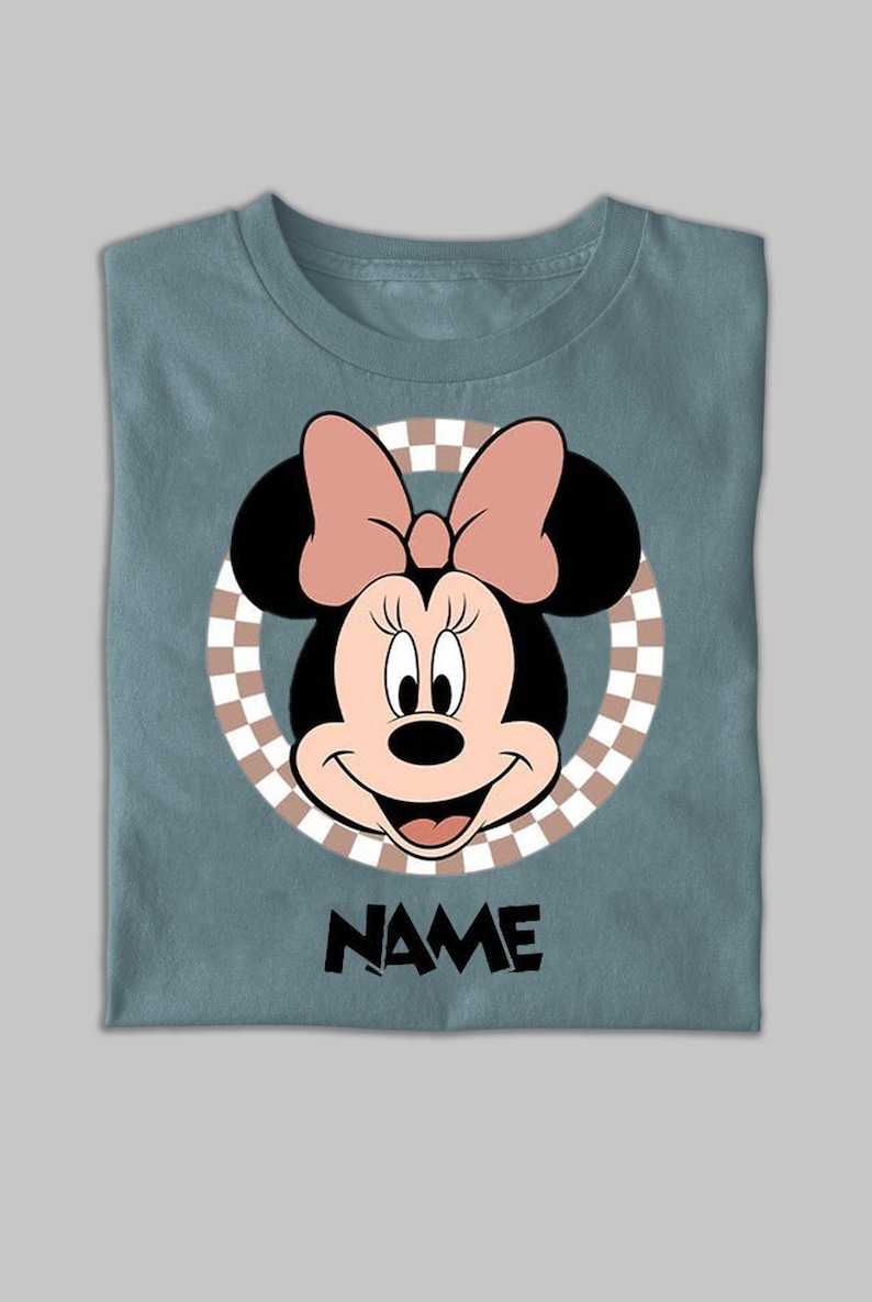 Puede incluir: Una camiseta azul pizarra con un gr&aacute;fico de Minnie Mouse centrado dentro de un c&iacute;rculo a cuadros beige y blanco. Minnie Mouse aparece con un lazo rosa, orejas negras y una sonrisa. La palabra NAME est&aacute; impresa en negro debajo.