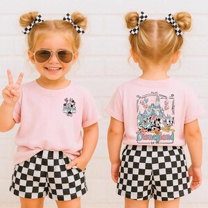 May include: A light pink t-shirt featuring a small graphic of a castle on the front left chest and a large graphic on the back depicting a castle with Disney characters and the text Disneyland Est. 1955, paired with black and white checkered shorts.