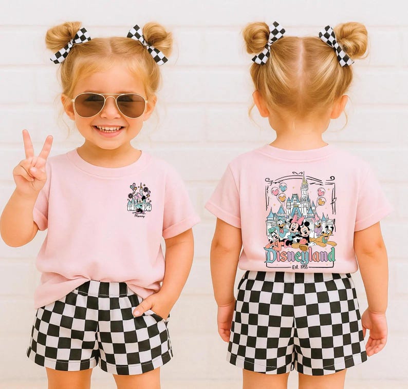 May include: A light pink t-shirt featuring a small graphic of a castle on the front left chest and a large graphic on the back depicting a castle with Disney characters and the text Disneyland Est. 1955, paired with black and white checkered shorts.