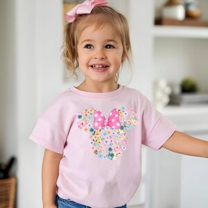 May include: A light pink short-sleeved t-shirt featuring a graphic of a mouse head silhouette composed of various colorful flowers and a large pink polka-dot bow centered at the top. The shirt has a crew neckline and a relaxed fit design.