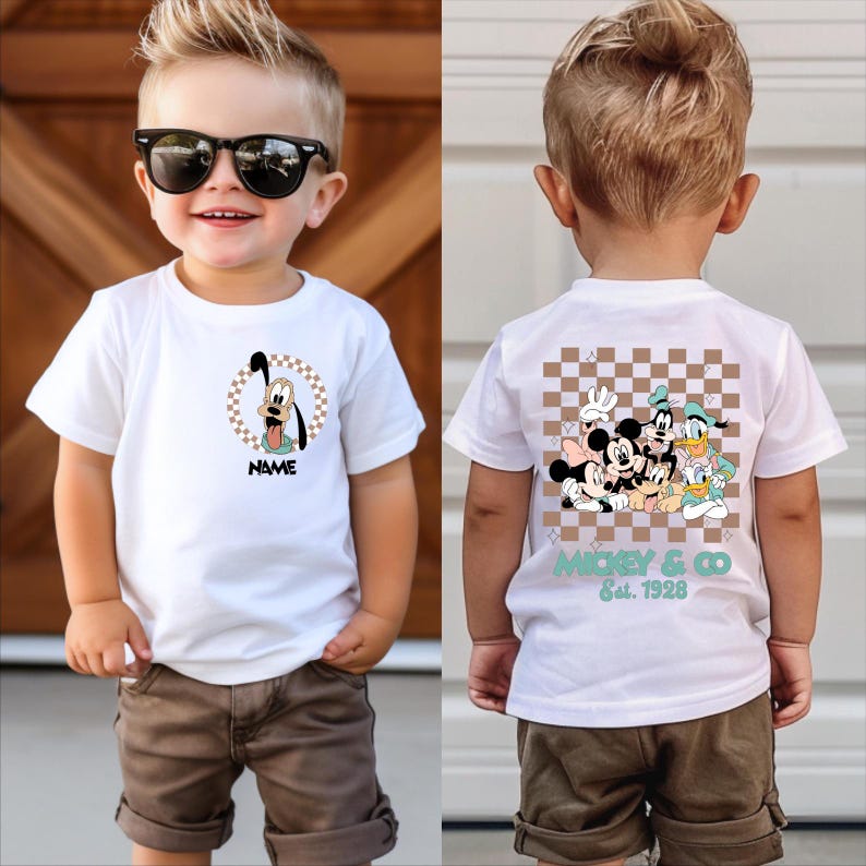 Puede incluir: Una camiseta blanca de manga corta con un gr&aacute;fico frontal de Goofy dentro de un c&iacute;rculo a cuadros y la palabra NAME debajo. La parte trasera muestra un gran cuadrado a cuadros con Mickey, Minnie, Goofy, Donald y Daisy, m&aacute;s MICKEY & CO Est. 1928.