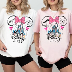 May include: A white t-shirt and a light pink t-shirt, each featuring a graphic of a castle, a pink polka-dot bow, Mickey Mouse ears, and the text "NAME", "Disney", and "2026". The designs include small pink balloons and firework accents.