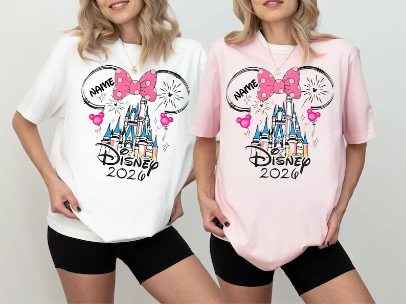 May include: A white t-shirt and a light pink t-shirt, each featuring a graphic of a castle, a pink polka-dot bow, Mickey Mouse ears, and the text "NAME", "Disney", and "2026". The designs include small pink balloons and firework accents.