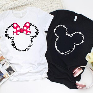 May include: A white t-shirt featuring a black outline of a mouse head shape filled with small mouse icons, a pink and white polka dot bow, and the text 2026, alongside a black t-shirt with a white mouse head outline and the text 2026.