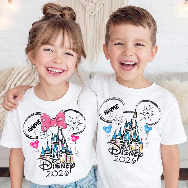 May include: Two white short-sleeved t-shirts featuring a graphic of a castle inside a Mickey Mouse ear outline. The design includes the text "NAME", "DISNEY 2026", fireworks, and small balloons. One shirt has a pink polka-dot bow on the ear.