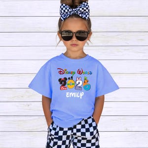 May include: A light blue t-shirt with the words "Disney World 2023" in colorful lettering. The shirt features cartoon character designs and the name "EMILY" in white. The child is wearing sunglasses and a black and white checkered bow.