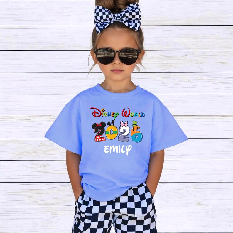May include: A light blue t-shirt with the words "Disney World 2023" in colorful lettering. The shirt features cartoon character designs and the name "EMILY" in white. The child is wearing sunglasses and a black and white checkered bow.