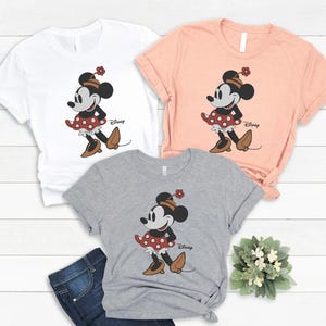 May include: Three short-sleeved crew neck t-shirts in white, peach, and heather grey. Each shirt features a graphic of Minnie Mouse wearing a polka dot skirt, a hat with a flower, and brown shoes, with the Disney logo printed in black script.