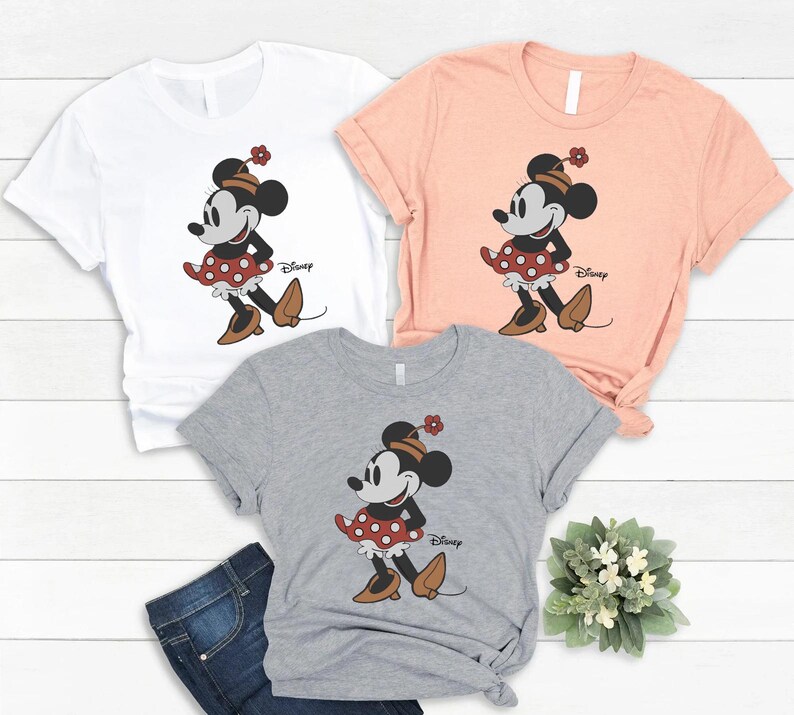 May include: Three short-sleeved crew neck t-shirts in white, peach, and heather grey. Each shirt features a graphic of Minnie Mouse wearing a polka dot skirt, a hat with a flower, and brown shoes, with the Disney logo printed in black script.