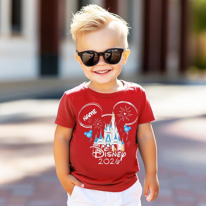 May include: A red short-sleeved t-shirt featuring a white graphic of a castle, fireworks, and Mickey Mouse ears. The text reads Name, Disney, and 2026. The design includes small blue balloons and white star accents on the front of the garment.