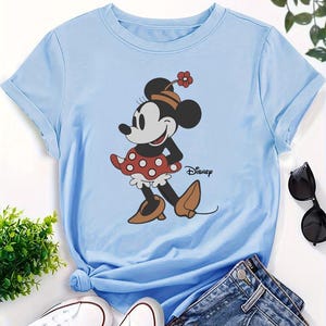 May include: A light blue short-sleeved t-shirt featuring a graphic of Minnie Mouse wearing a red and white polka dot skirt, a hat with a small flower, and brown shoes. The word Disney is printed in black script next to the character illustration.