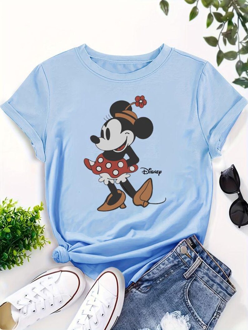 May include: A light blue short-sleeved t-shirt featuring a graphic of Minnie Mouse wearing a red and white polka dot skirt, a hat with a small flower, and brown shoes. The word Disney is printed in black script next to the character illustration.