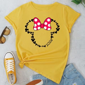 May include: A bright yellow short-sleeved t-shirt featuring a graphic of a Minnie Mouse-style bow in red with white polka dots, outlined by a silhouette of small black Mickey Mouse heads and the text 2020 printed in black on the lower right side.