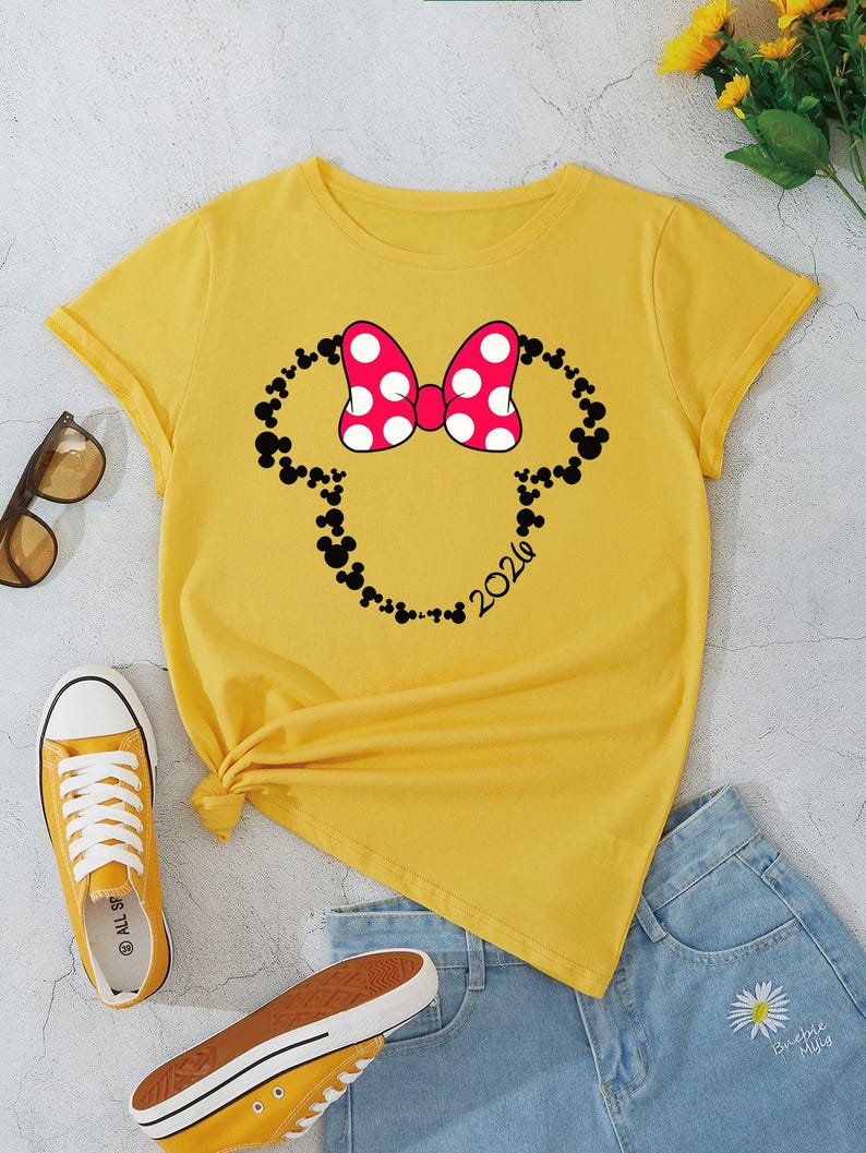 May include: A bright yellow short-sleeved t-shirt featuring a graphic of a Minnie Mouse-style bow in red with white polka dots, outlined by a silhouette of small black Mickey Mouse heads and the text 2020 printed in black on the lower right side.