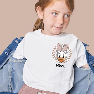 Puede incluir: Una camiseta blanca de manga corta con un gr&aacute;fico circular en el pecho. El gr&aacute;fico muestra un personaje de pato de dibujos animados con un lazo rosa, sobre un fondo de cuadros beige y blanco, con la palabra NAME impresa en negro debajo.