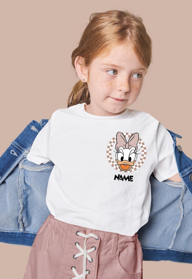 Puede incluir: Una camiseta blanca de manga corta con un gr&aacute;fico circular en el pecho. El gr&aacute;fico muestra un personaje de pato de dibujos animados con un lazo rosa, sobre un fondo de cuadros beige y blanco, con la palabra NAME impresa en negro debajo.