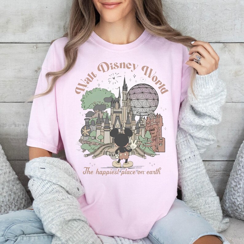May include: A light pink t-shirt featuring a graphic of Walt Disney World with Mickey Mouse. The design includes a castle, Spaceship Earth, and the text "Walt Disney World" and "The happiest place on earth."
