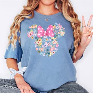 May include: A blue short-sleeved t-shirt featuring a graphic of a Mickey Mouse head silhouette composed of various colorful flowers. A large pink bow with white polka dots is positioned at the top center of the floral mouse head design.