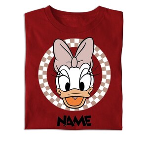 Puede incluir: Una camiseta de color rojo oscuro con un gr&aacute;fico de Daisy Duck con un lazo rosa claro. El personaje est&aacute; centrado dentro de un marco circular con un patr&oacute;n de cuadros marrones y blancos. Debajo del gr&aacute;fico, la palabra NAME est&aacute; en letras negras.