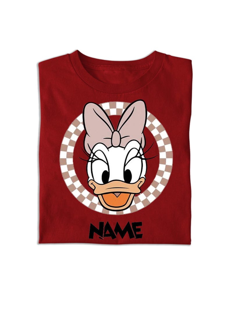Puede incluir: Una camiseta de color rojo oscuro con un gr&aacute;fico de Daisy Duck con un lazo rosa claro. El personaje est&aacute; centrado dentro de un marco circular con un patr&oacute;n de cuadros marrones y blancos. Debajo del gr&aacute;fico, la palabra NAME est&aacute; en letras negras.