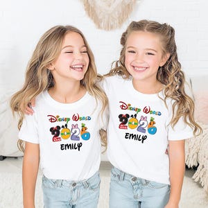 May include: Two young girls wearing white t-shirts with a colorful "Disney World 2020" graphic and the name "Emily" printed below. The shirts feature Mickey and Minnie Mouse, a bow, and a pumpkin. The girls are smiling.