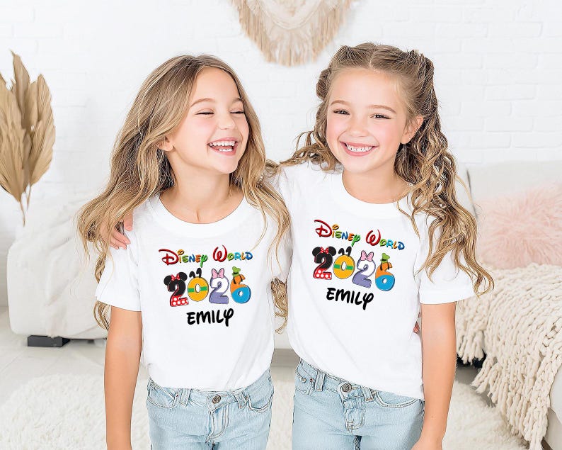 May include: Two young girls wearing white t-shirts with a colorful "Disney World 2020" graphic and the name "Emily" printed below. The shirts feature Mickey and Minnie Mouse, a bow, and a pumpkin. The girls are smiling.