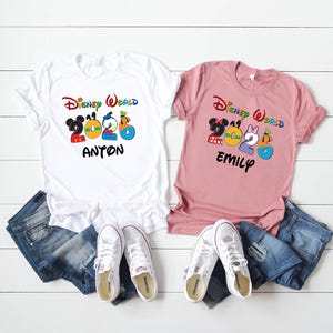 May include: Two t-shirts with the text "Disney World 2020" and names. One white shirt has "Anton" and the other is pink with "Emily". Both shirts have colorful cartoon character graphics. Blue jeans and white sneakers are also in the image.