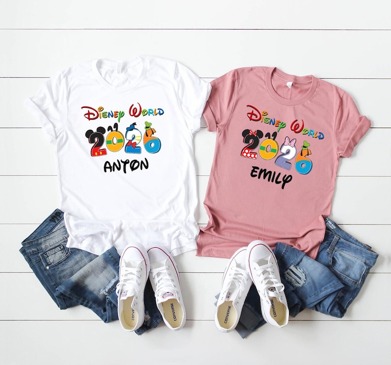May include: Two t-shirts with the text "Disney World 2020" and names. One white shirt has "Anton" and the other is pink with "Emily". Both shirts have colorful cartoon character graphics. Blue jeans and white sneakers are also in the image.