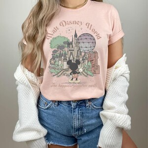 May include: A light pink t-shirt featuring a graphic of Walt Disney World with Mickey Mouse. The design includes Cinderella's castle, Spaceship Earth, and the text "Walt Disney World" and "The happiest place on earth."