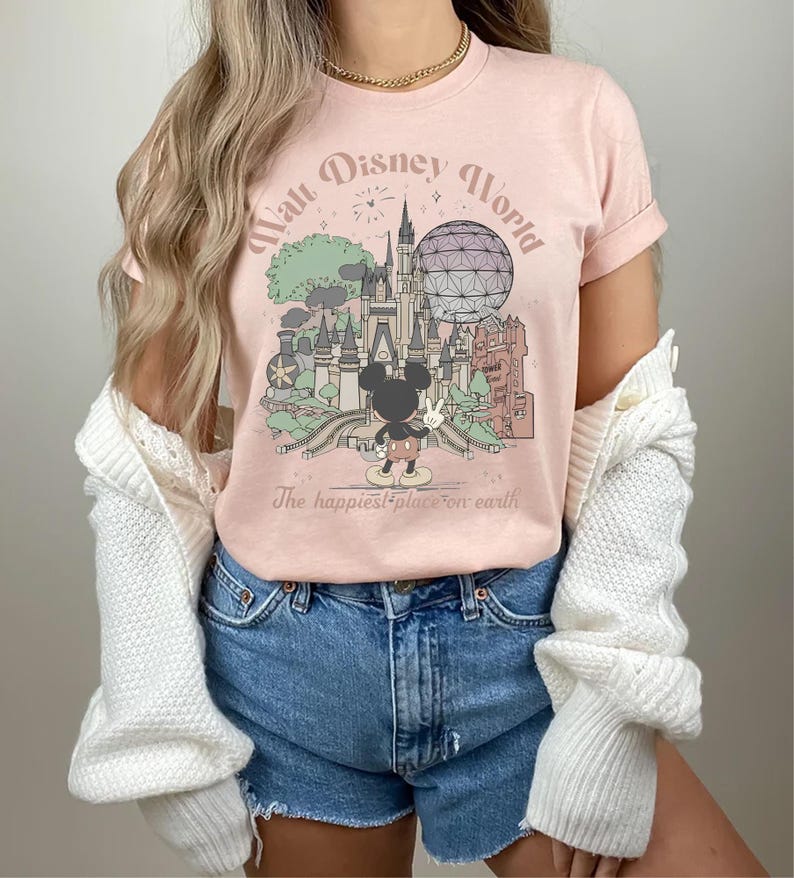 May include: A light pink t-shirt featuring a graphic of Walt Disney World with Mickey Mouse. The design includes Cinderella's castle, Spaceship Earth, and the text "Walt Disney World" and "The happiest place on earth."