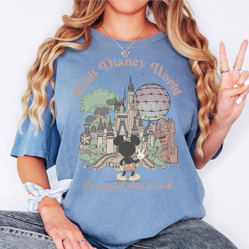 May include: Blue t-shirt with a vintage-style Walt Disney World graphic. The design features Cinderella's castle, Spaceship Earth, and Mickey Mouse. Text on the shirt reads "Walt Disney World" and "The happiest place on earth."