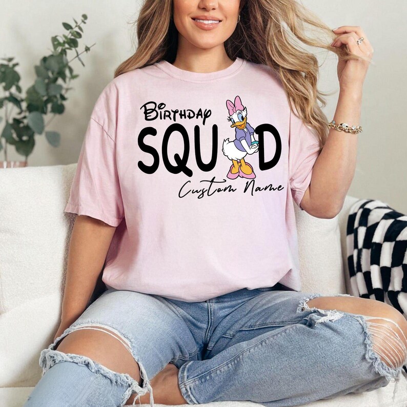 Pode incluir: Uma t-shirt de manga curta cor-de-rosa claro com o texto preto "Birthday SQUAD", onde a letra "A" &eacute; substitu&iacute;da por um gr&aacute;fico da Daisy Duck. Abaixo do texto, as palavras "Custom Name" est&atilde;o impressas numa fonte cursiva preta.