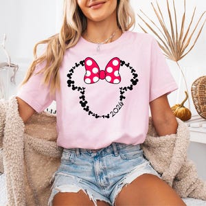 May include: A light pink short-sleeved t-shirt featuring a graphic of a mouse-ear silhouette outlined in small black mouse-ear icons. A large pink bow with white polka dots sits at the top, and the number 2026 is printed in black script.