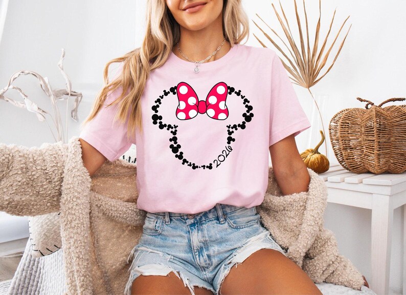 May include: A light pink short-sleeved t-shirt featuring a graphic of a mouse-ear silhouette outlined in small black mouse-ear icons. A large pink bow with white polka dots sits at the top, and the number 2026 is printed in black script.