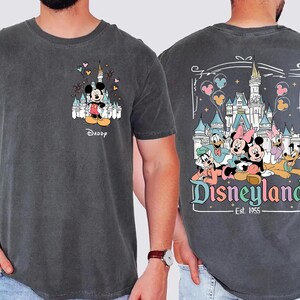 May include: A charcoal gray t-shirt featuring a small graphic of Mickey Mouse and a castle on the front chest with the word Daddy below it. The back displays a large graphic of Mickey, Minnie, Donald, Daisy, Goofy, and Pluto at Disneyland.